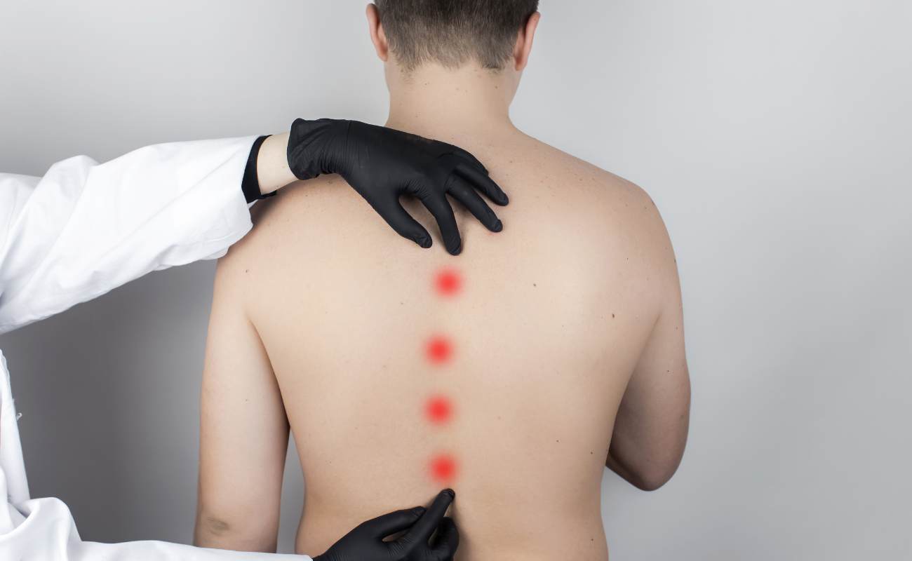 Na zdjęciu widzimy osobę w białym lekarskim płaszczu z czarnymi lateksowymi rękawiczkami, która wykonuje badanie kręgosłupa piersiowego na pacjencie. Pacjent ma odsłonięte plecy z widocznymi pięcioma czerwonymi kropkami umieszczonymi wzdłuż kręgosłupa, które mogą oznaczać miejsca do akupunktury lub punkty orientacyjne dla badania. Tło jest jednolite i jasne.