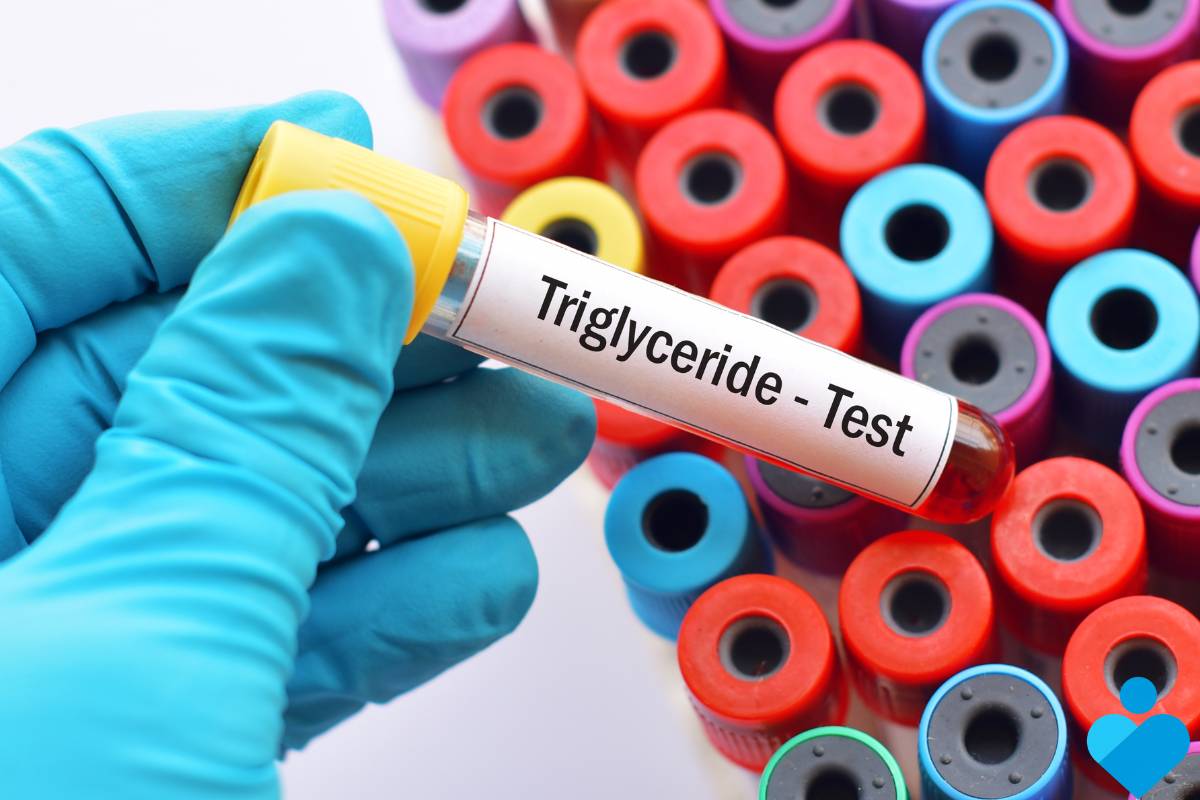 Na zdjęciu widzimy rękę w niebieskiej lateksowej rękawiczce trzymającą probówkę z krwią oznaczoną jako "Triglyceride - Test". Tło zdjęcia jest rozmyte, ale widoczne są liczne zakrętki probówek w różnych kolorach, co sugeruje, że zdjęcie zostało wykonane w laboratorium medycznym. Etap ten wskazuje na przeprowadzenie specyficznego testu krwi mającego na celu pomiar poziomu trójglicerydów w organizmie.