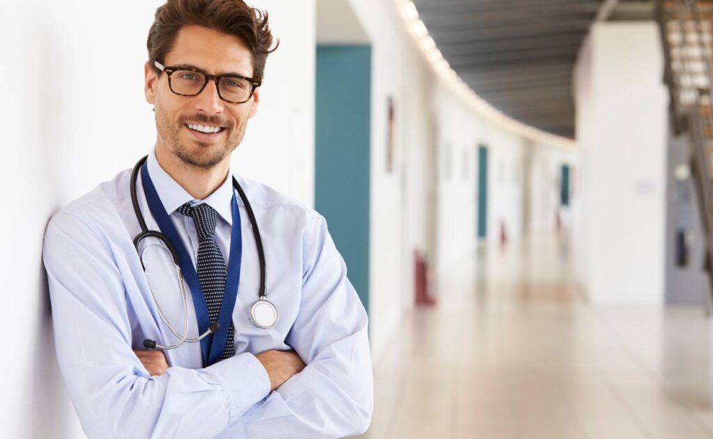 Na zdjęciu widzimy mężczyznę w białym lekarskim fartuchu z założonym stetoskopem na szyi. Ma na sobie niebieską koszulę, krawat w drobne wzory i okulary. Uśmiecha się i stoi z założonymi rękami, co sugeruje pewność siebie i przyjazną postawę. Tło jest lekko rozmyte, ale można zauważyć długi korytarz szpitala lub kliniki. Mężczyzna wydaje się być lekarzem rodzinnym lub specjalistą w środowisku medycznym.