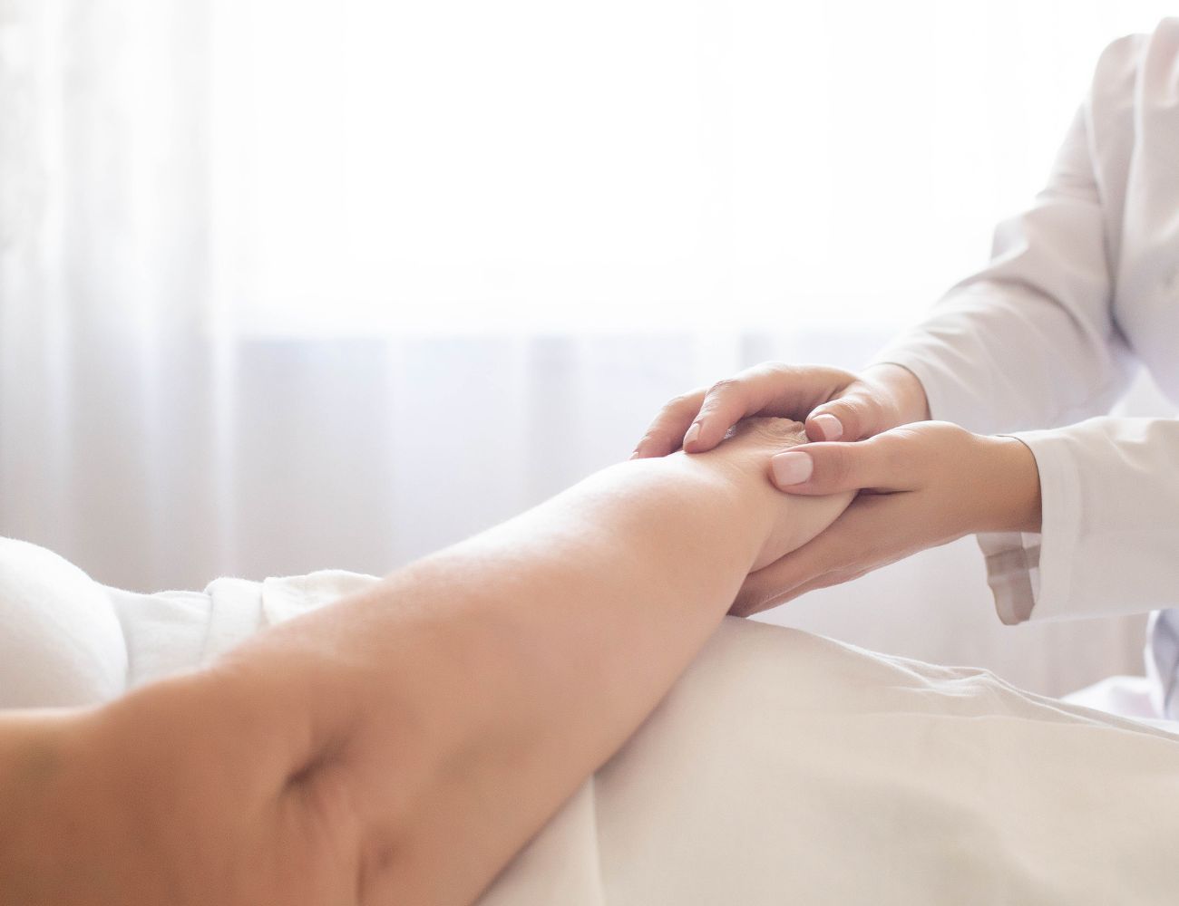 Zdjęcie główna w artykule "Babcine sposoby na odleżyny" Zdjęcie pokazuje zbliżenie na dłonie osoby ubranej w biały uniform medyczny, która delikatnie obejmuje rękę pacjenta leżącego na łóżku. Sceneria jest jasna i spokojna, sugerując pielęgnację lub badanie medyczne. Całość oddaje atmosferę troski i profesjonalizmu w kontekście medycznym lub opiekuńczym.