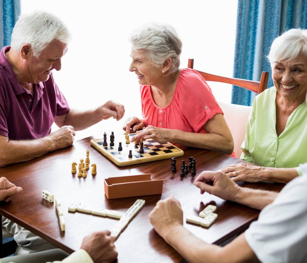 Zdjęcie przedstawia czterech starszych ludzi grających w różne gry planszowe i towarzyskie. Na pierwszym planie widzimy starszego mężczyznę w bordowej koszulce i starszą kobietę w czerwonej bluzce, którzy grają w szachy. Mężczyzna uśmiecha się, przenosząc szachową figurę. Po prawej stronie kadru siedzi starsza kobieta w zielonej bluzce, która uśmiecha się, obserwując grę. W tle widoczna jest jeszcze jedna kobieta, biorąca udział w grze w domino. Wszyscy znajdują się w jasnym pomieszczeniu z widokiem na zasłonięte okno, a atmosfera jest radosna i pełna przyjaznej rywalizacji.