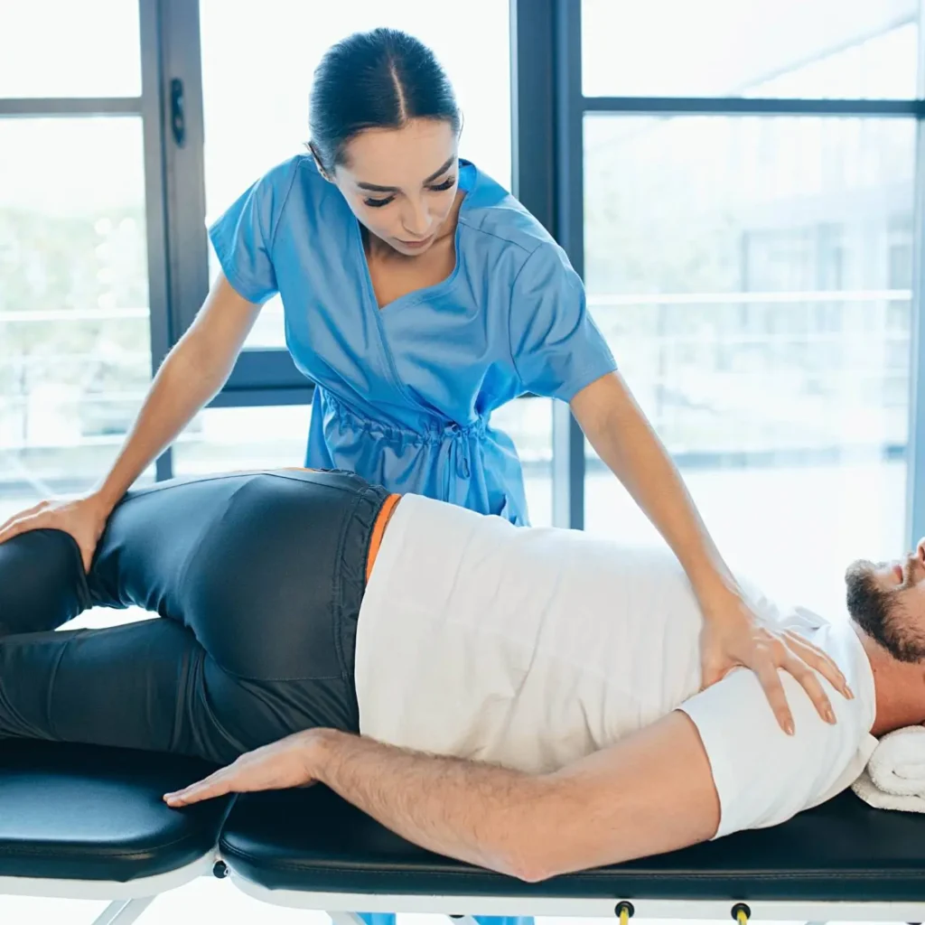Zdjęcie przedstawia scenę z sesji fizjoterapeutycznej. Fizjoterapeutka ubrana w niebieski uniform przeprowadza zabieg na mężczyźnie leżącym na boku na specjalistycznym stole. Kobieta używa rąk, aby manipulować ciałem pacjenta, skupiając się na jego plecach. Mężczyzna ma na sobie białą koszulkę i ciemne spodnie. Otoczenie sugeruje, że sesja odbywa się w jasnym, nowoczesnym gabinecie z dużymi oknami, przez które wpada naturalne światło.