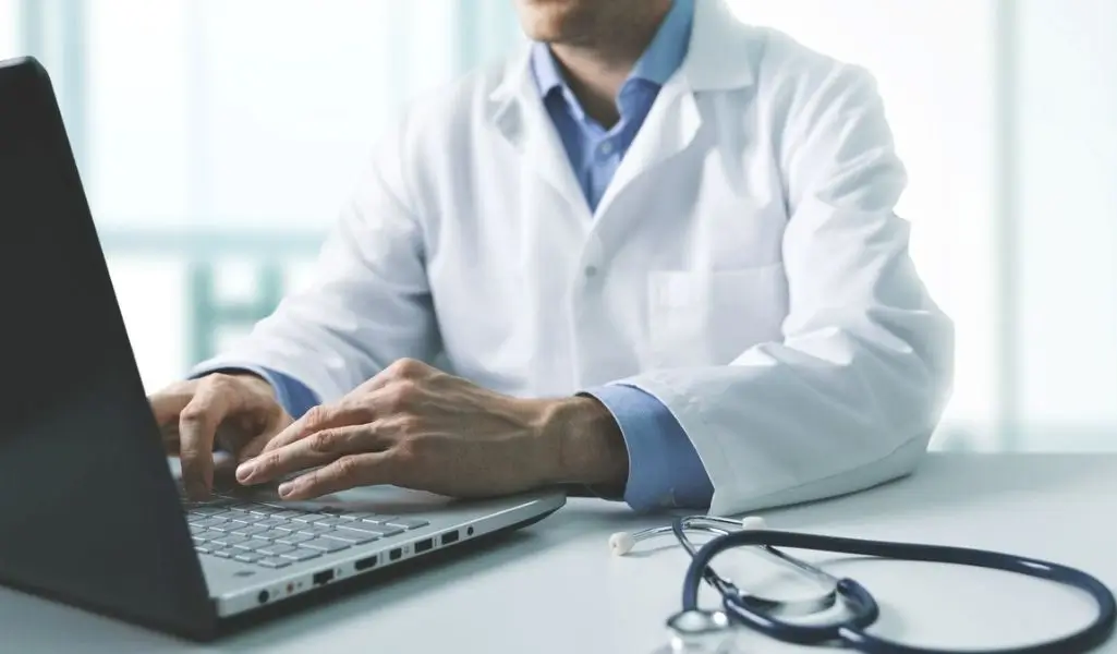 Obrazek wyróżniający do artykułu - zwolnienie lekarskie wstecz. Na zdjęciu widzimy lekarza siedzącego przy biurku, który pracuje na laptopie. Lekarz ubrany jest w biały kitel i niebieską koszulę. Na biurku, obok laptopa, leży stetoskop, co dodatkowo sugeruje, że jest to osoba związana z medycyną. Tło zdjęcia jest rozmyte, ale widoczny jest jasny, prawdopodobnie dobrze oświetlony pokój, co nadaje fotografii profesjonalny i sterylny wygląd typowy dla środowiska medycznego.