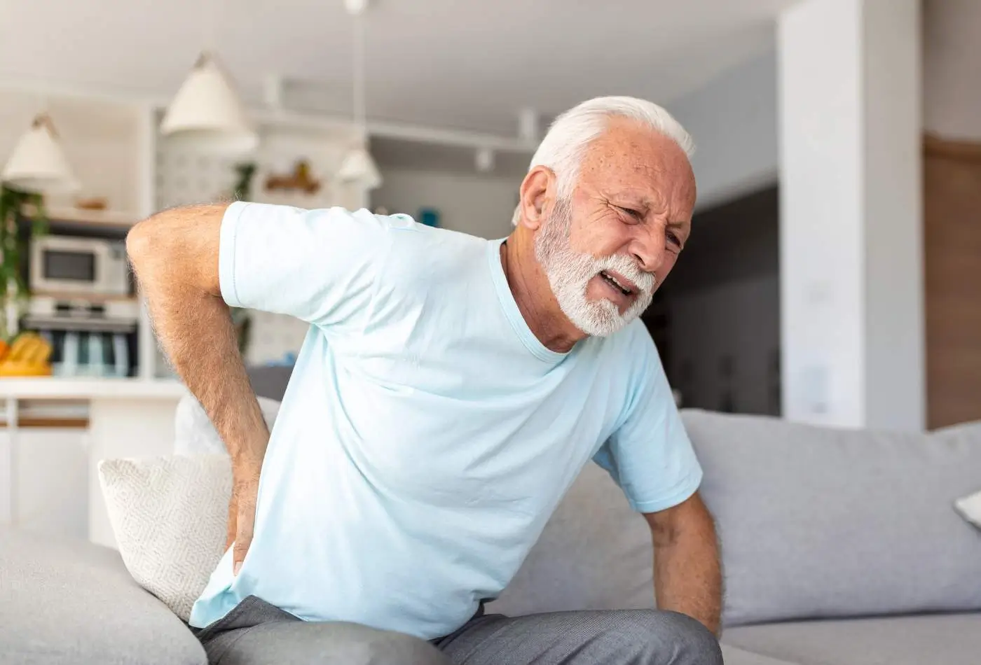 Obraz przedstawia starszego mężczyznę, który siedzi na kanapie w jasnym, nowoczesnym wnętrzu. Mężczyzna ma siwe włosy i brodę, ubrany jest w jasnoniebieską koszulkę. Wyraz jego twarzy wskazuje na ból lub dyskomfort. Trzyma się za dolną część pleców, co sugeruje, że może odczuwać ból krzyża lub rwy kulszowej. W tle widoczne są elementy kuchni oraz dekoracje domowe, takie jak rośliny i lampy wiszące.