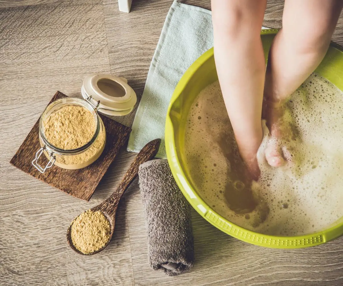 Obraz przedstawia scenę związaną z domowym zabiegiem na poprawę krążenia w nogach. ### Opis szczegółowy: 1. **Nogi w misce**: - Dwie nogi zanurzone w okrągłej, jasnozielonej misce z wodą. Woda jest spieniona, co sugeruje, że dodano do niej jakiś środek mydlany lub relaksacyjny. 2. **Słoik z proszkiem**: - Obok miski znajduje się otwarty słoik ze szczelnym, zatrzaskowym zamknięciem. Słoik jest wypełniony żółtawym proszkiem, który może być solą do kąpieli lub innym preparatem do kąpieli stóp. 3. **Łyżka i proszek**: - Drewniana łyżka leży na podłodze, a w niej również znajduje się trochę tego samego proszku, co w słoiku. Łyżka sugeruje, że proszek został dozowany ręcznie. 4. **Ręczniki**: - Na podłodze w pobliżu miski znajduje się szary, złożony ręcznik oraz jasnozielona ściereczka. 5. **Podłoga**: - Cała scena jest ustawiona na podłodze o jasnym, drewnianym wykończeniu, co nadaje naturalnego i przytulnego wyglądu. ### Ogólny wygląd: - Obraz emanuje spokojem i domową atmosferą. Wyraźnie sugeruje zabieg relaksacyjny, który ma na celu poprawę krążenia w nogach lub relaksację stóp. - Kolory są stonowane, a całość aranżacji jest dobrze skomponowana i estetyczna. Obraz doskonale oddaje ideę domowego spa lub zabiegu pielęgnacyjnego, skoncentrowanego na poprawie samopoczucia i zdrowia stóp.