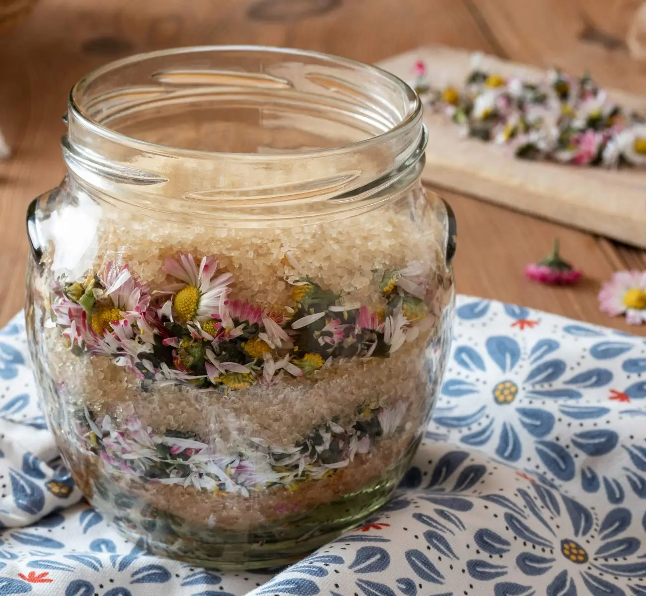 Obraz przedstawia słoik z warstwowym układem cukru i stokrotek, umieszczony na stole. Słoik jest wypełniony na przemian warstwami brązowego cukru i kwiatów stokrotek, co tworzy atrakcyjny wizualnie efekt. Tło stanowi drewniany stół, na którym leży słoik oraz kilka luźnych stokrotek. Słoik stoi na białej ściereczce z niebieskim, kwiatowym wzorem, co dodaje uroku i podkreśla rustykalny klimat.