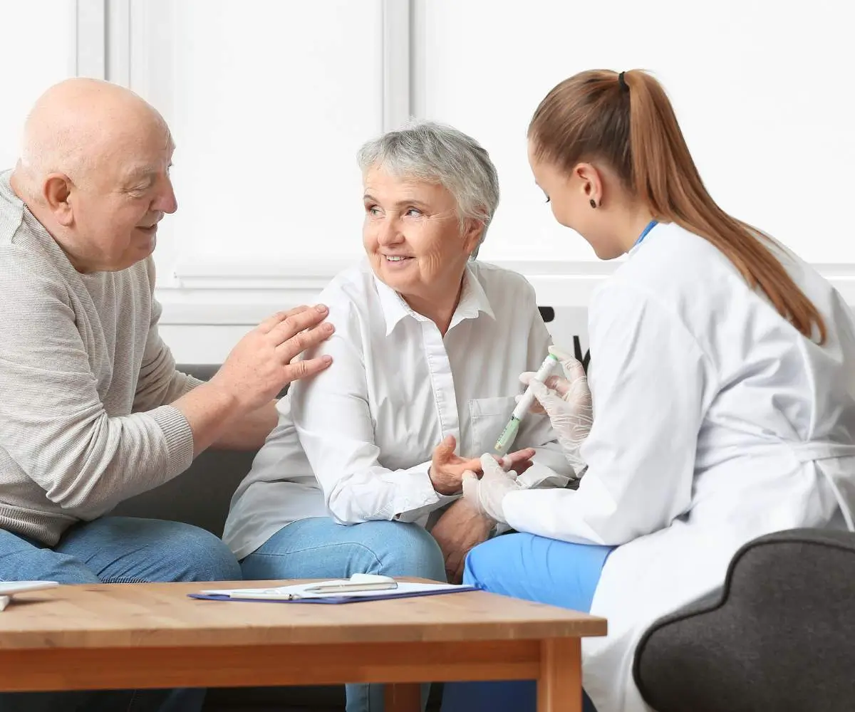 Na zdjęciu widoczna jest scena w pomieszczeniu, gdzie starsza kobieta i starszy mężczyzna siedzą na kanapie, rozmawiając z młodą kobietą w białym kitlu, która prawdopodobnie jest lekarzem lub pielęgniarką. Starsza kobieta ma krótkie, siwe włosy i jest ubrana w białą koszulę. Mężczyzna ma łysą głowę i jest ubrany w jasnoszary sweter. Młoda kobieta w kitlu trzyma w ręku igłę do zastrzyków i zakłada rękawiczki, co sugeruje, że zamierza podać zastrzyk lub pobrać krew. Atmosfera jest spokojna i przyjazna, a starsza kobieta uśmiecha się w stronę mężczyzny, który z nią rozmawia. Tło jest jasne, co sugeruje dobrze oświetlone, przytulne wnętrze.