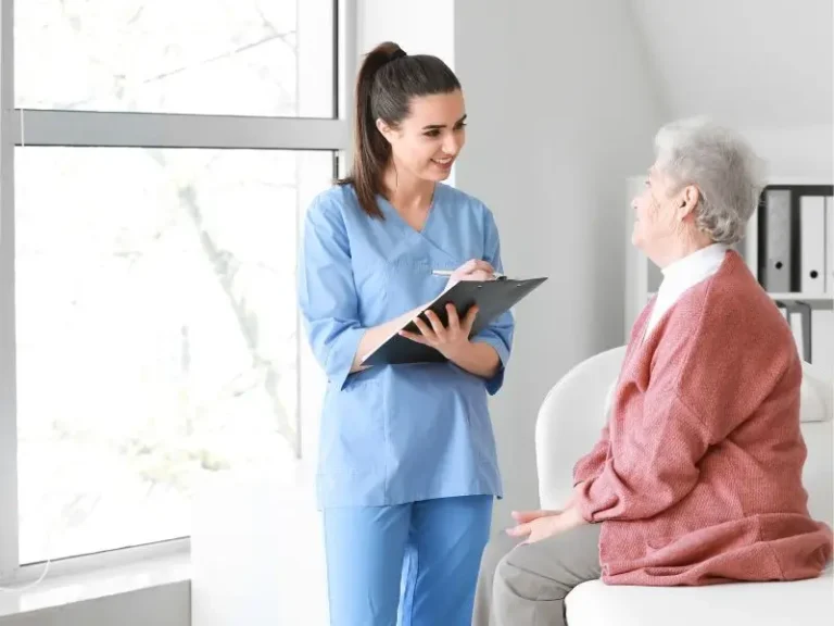Na zdjęciu widzimy młodą kobietę ubraną w niebieski strój medyczny, prawdopodobnie pielęgniarkę lub opiekunkę, rozmawiającą z starszą kobietą. Starsza kobieta siedzi na krześle ubrana w różowy sweter, wygląda spokojnie i uśmiecha się. Kobieta w stroju medycznym trzyma w rękach clipboard z dokumentami, na które najprawdopodobniej coś zapisuje. Scena sugeruje spokojną, przyjazną rozmowę, być może dotyczącą opieki zdrowotnej. Tło to jasne pomieszczenie z dużym oknem, przez które wpada naturalne światło, co dodaje zdjęciu jasnej i pozytywnej atmosfery.