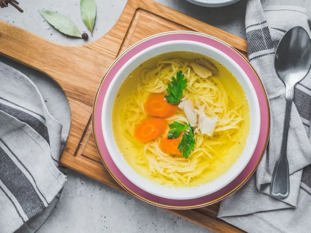 Na zdjęciu znajduje się miska z rosołem podana na drewnianej desce. W środku widać jasny bulion z makaronem, plasterkami gotowanej marchewki, kawałkami kurczaka oraz liśćmi natki pietruszki, które nadają daniu świeżości. Obok miski leży metalowa łyżka i serwetka w odcieniach szarości z wzorem w kratkę. Na desce widoczne są również dwa liście laurowe i kilka przypraw. Całość prezentuje się estetycznie i apetycznie.