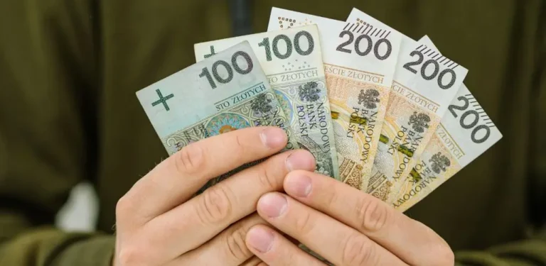 Obrazek wyróżniający do artykułu - ile pieniędzy przysługuje osobie z grupą. Na obrazie widzimy ręce trzymające wachlarz z banknotów polskich o nominałach 100 i 200 złotych. Banknoty są ułożone w wachlarz, a dłonie je przytrzymujące są lekko zaciśnięte. Banknoty mają charakterystyczne kolory: 100 złotych to odcienie zieleni, a 200 złotych to odcienie złota i brązu. Tło jest rozmyte, co sprawia, że uwaga skupiona jest na pieniądzach.