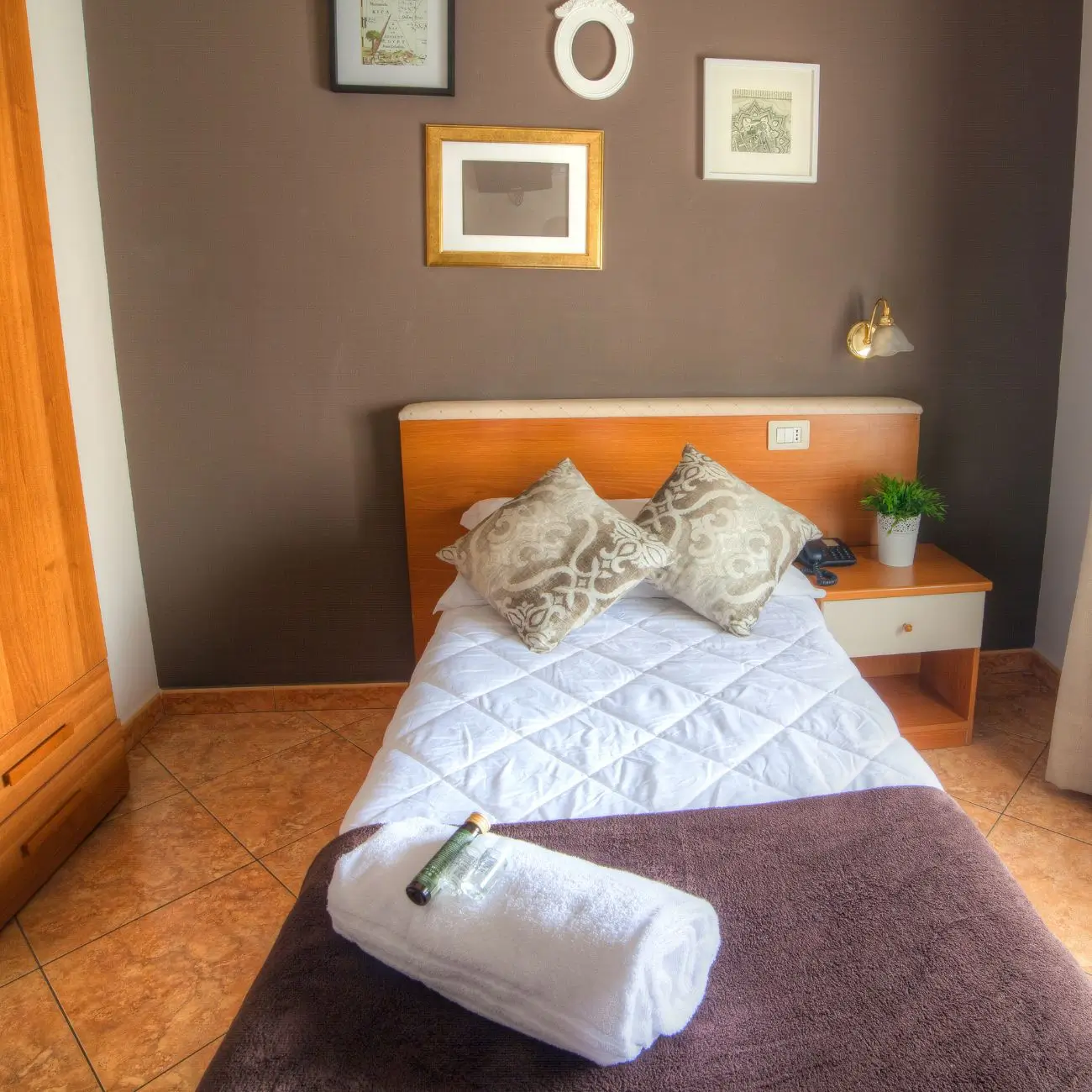 Obraz przedstawia fragment pokoju z jednoosobowym łóżkiem nakrytym narzutą i dwoma ozdobnymi poduszkami w kolorach brązowym i białym. Nad zagłówkiem znajduje się drewniana półka z lampką oraz kilkoma ozdobnymi ramkami ze zdjęciami i obrazami na ścianie pomalowanej na ciemny kolor. Na podłodze z płytek ułożona jest brązowa wykładzina. Całość tworzy przytulne i zadbane wnętrze sypialni utrzymane w stonowanych barwach, sprzyjające odpoczynkowi.