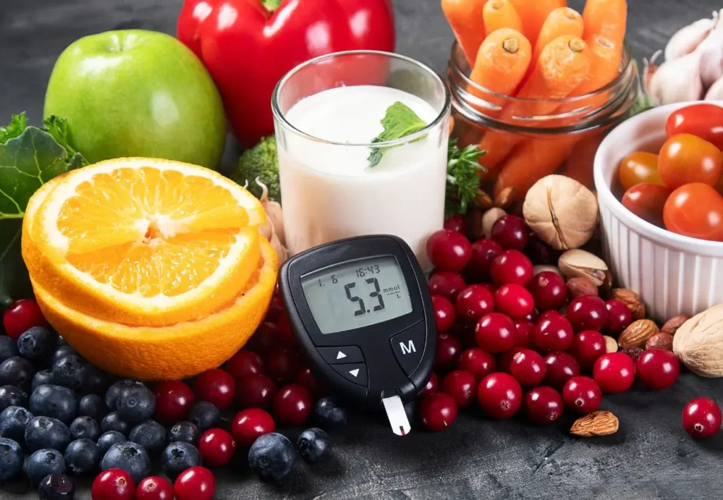 Na zdjęciu widoczny jest zestaw zdrowej żywności, która może być korzystna dla osób z cukrzycą. W centrum zdjęcia znajduje się glukometr pokazujący poziom cukru wynoszący 5,3 mmol/L. Obok niego ułożone są różnorodne owoce i warzywa, takie jak: • Połówki pomarańczy, • Jabłko (zielone), • Papryka czerwona, • Brokuły, • Borówki, żurawina i wiśnie, • Pomidorki koktajlowe, • Marchewki (w szklanym słoiku), • Orzechy i migdały w miseczce. Obok znajduje się także szklanka mleka z listkiem mięty na wierzchu.