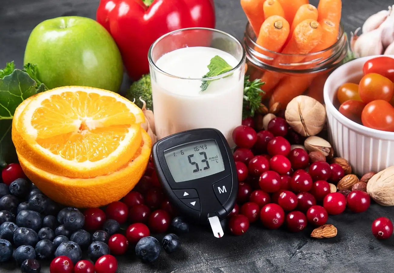 Na zdjęciu widoczny jest zestaw zdrowej żywności, która może być korzystna dla osób z cukrzycą. W centrum zdjęcia znajduje się glukometr pokazujący poziom cukru wynoszący 5,3 mmol/L. Obok niego ułożone są różnorodne owoce i warzywa, takie jak: • Połówki pomarańczy, • Jabłko (zielone), • Papryka czerwona, • Brokuły, • Borówki, żurawina i wiśnie, • Pomidorki koktajlowe, • Marchewki (w szklanym słoiku), • Orzechy i migdały w miseczce. Obok znajduje się także szklanka mleka z listkiem mięty na wierzchu.