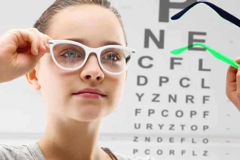 Obraz przedstawia młodą dziewczynę zakładającą białe okulary korekcyjne. W tle widoczna jest tablica okulistyczna z literami różnej wielkości, używana do testów ostrości wzroku. Po prawej stronie widoczna jest dłoń trzymająca kolorowe oprawki, co sugeruje wybór odpowiednich okularów podczas badania wzroku.