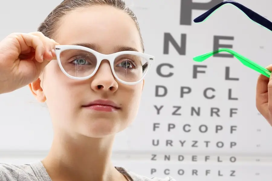 Obraz przedstawia młodą dziewczynę zakładającą białe okulary korekcyjne. W tle widoczna jest tablica okulistyczna z literami różnej wielkości, używana do testów ostrości wzroku. Po prawej stronie widoczna jest dłoń trzymająca kolorowe oprawki, co sugeruje wybór odpowiednich okularów podczas badania wzroku.