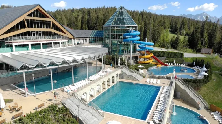 Na zdjęciu widoczny jest kompleks basenów termalnych na tle górskiego krajobrazu. Architektura obiektu łączy nowoczesne rozwiązania, takie jak przeszklona piramida i zadaszenia nad basenami, z elementami nawiązującymi do tradycyjnych stylów, jak skośne dachy budynków. Kompleks składa się z kilku basenów, zarówno krytych, jak i odkrytych, otoczonych białymi leżakami. Z lewej strony widać miejsca do wypoczynku z parasolami i stolikami. W centralnej części zdjęcia wyróżnia się mostek z arkadami, łączący różne sekcje basenowe. Po prawej stronie zauważalne są kolorowe zjeżdżalnie wodne w odcieniach niebieskim, żółtym i czerwonym, prowadzące do basenu rekreacyjnego. Otoczenie kompleksu to zielone tereny, las i góry widoczne w tle, co podkreśla malowniczy charakter tego miejsca. Całość tworzy przyjemną atmosferę wypoczynku i relaksu.