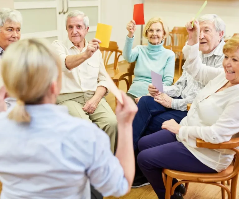 Zdjęcie przedstawia grupę seniorów siedzących w półokręgu na drewnianych krzesłach w jasnym wnętrzu. Uczestniczą oni w zajęciach grupowych, podczas których każdy z nich trzyma w ręku kolorową kartkę – żółtą, czerwoną, fioletową lub zieloną. Wyglądają na zaangażowanych i zadowolonych, a ich postawy sugerują aktywne uczestnictwo w ćwiczeniu lub zabawie. Na pierwszym planie, z tyłu, widoczna jest osoba prowadząca zajęcia, ubrana w jasnoniebieską koszulę. Atmosfera na zdjęciu jest ciepła i radosna, co sugeruje wspierającą i aktywizującą formę spędzania czasu dla seniorów.