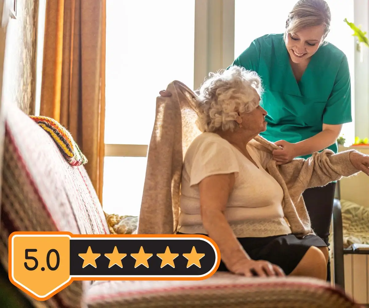 Zdjęcie przedstawia scenę w jasnym wnętrzu, w której młodsza kobieta w zielonym uniformie, sugerującym pracę w opiece medycznej lub społecznej, pomaga starszej kobiecie założyć sweter. Starsza kobieta siedzi na kanapie z kolorowym kocem w tle. Wygląda na zadowoloną, a opiekunka uśmiecha się, co tworzy ciepłą i troskliwą atmosferę. W lewym dolnym rogu zdjęcia znajduje się graficzne oznaczenie z oceną “5.0” oraz pięcioma złotymi gwiazdkami, co może sugerować wysoką jakość usług lub opinię na temat placówki lub osoby przedstawionej na zdjęciu.