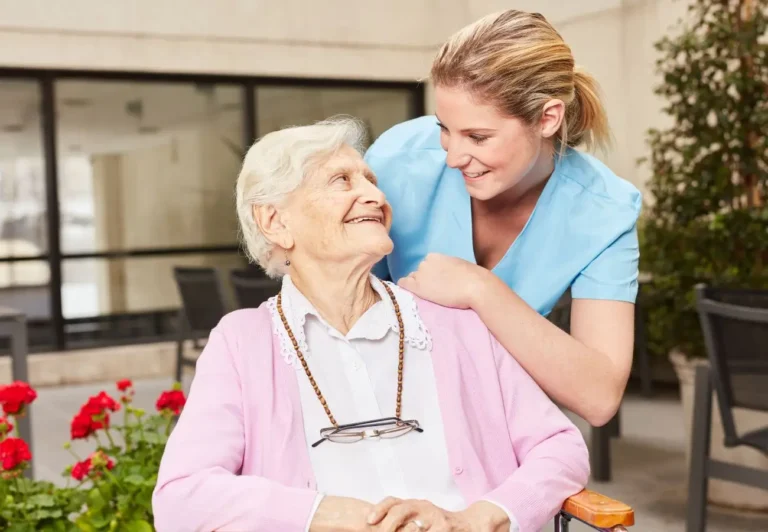Obraz przedstawia młodą opiekunkę medyczną w niebieskim stroju, pochyloną z uśmiechem nad starszą kobietą siedzącą na wózku inwalidzkim. Starsza kobieta w różowym swetrze i z zawieszonym na szyi różańcem również uśmiecha się, patrząc na opiekunkę. Scena ma miejsce na zewnątrz, w otoczeniu roślin i czerwonych kwiatów, co podkreśla pogodną i serdeczną atmosferę.
