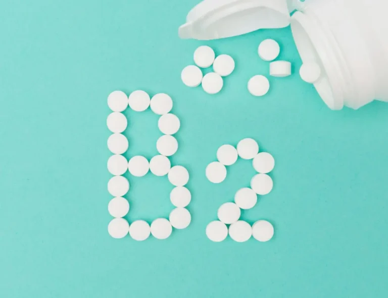 Zdjęcie przedstawia kompozycję, na której białe tabletki ułożone są w kształt liter „B2”, symbolizujących witaminę B2 (ryboflawinę). Tło jest jasnoturkusowe, co nadaje fotografii czysty i minimalistyczny wygląd. Po prawej stronie widoczna jest biała, otwarta butelka, z której wysypują się tabletki, tworząc dodatkowy element wizualny. Kompozycja podkreśla temat suplementów diety lub zdrowia.