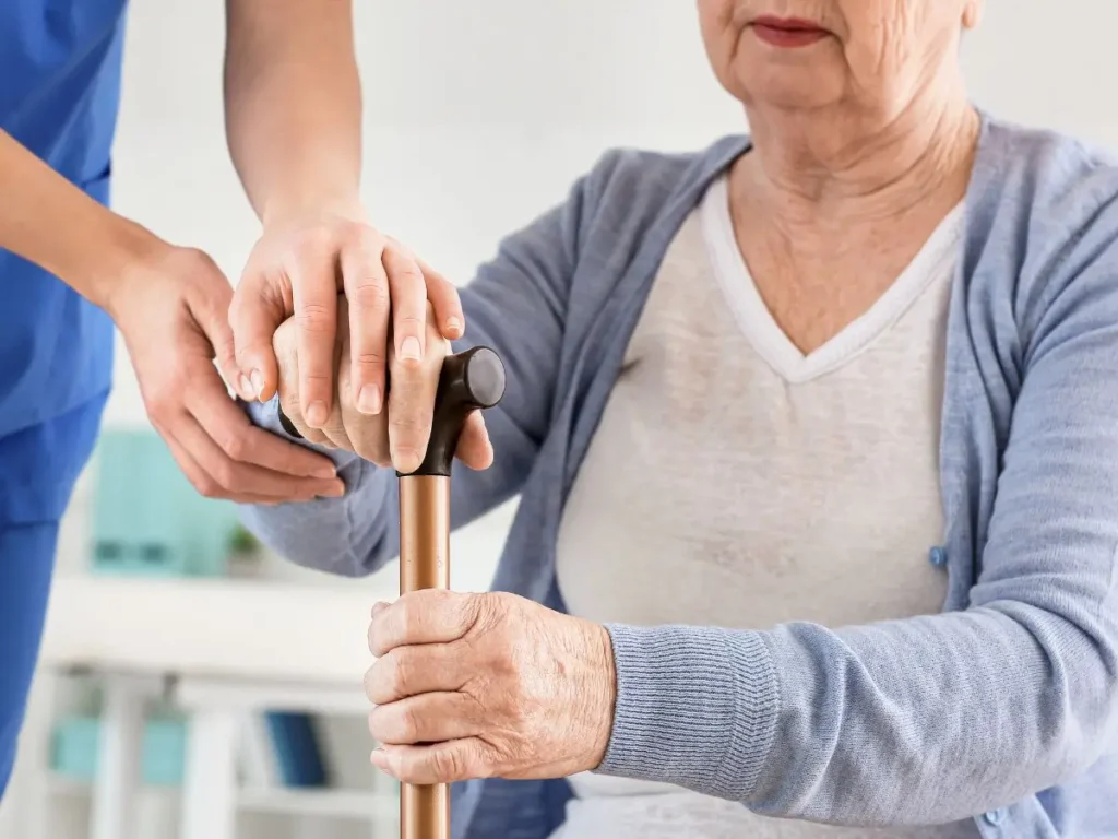 Na zdjęciu widzimy starszą kobietę, której ręce spoczywają na uchwycie laski. Kobieta jest ubrana w białą koszulkę i jasnoniebieski sweter. Jej dłonie są widoczne na pierwszym planie – trzymają laskę w sposób pewny, choć świadczący o potrzebie wsparcia. Obok znajduje się osoba w niebieskim uniformie, prawdopodobnie pielęgniarka lub opiekun, która delikatnie kładzie swoją rękę na dłoni starszej kobiety, okazując wsparcie i troskę. Gest pielęgniarki wyraża ciepło i chęć pomocy, a jednocześnie podkreśla opiekuńczy charakter sytuacji. Tło jest jasne i rozmyte, co sugeruje wnętrze medyczne lub opiekuńcze, takie jak dom opieki, szpital lub prywatne mieszkanie. Zdjęcie emanuje spokojem, troską i atmosferą wsparcia, ukazując relację między osobą potrzebującą pomocy a jej opiekunem.