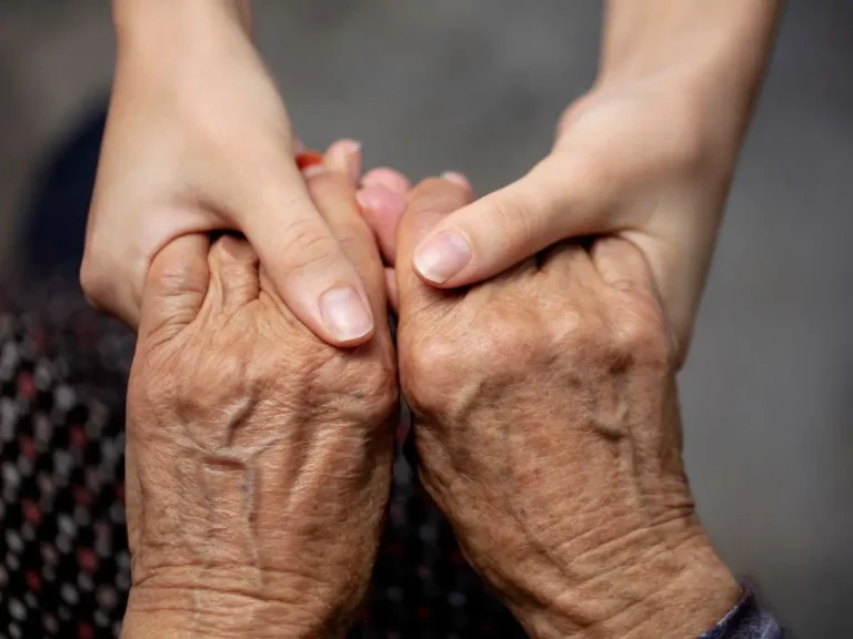 Zdjęcie przedstawia symboliczną scenę wyrażającą wsparcie, troskę i międzypokoleniowe więzi. Widoczne są dwie pary rąk: starszej osoby i młodszej. Starsza osoba ma dłonie widoczne na pierwszym planie – ich skóra jest pomarszczona, z widocznymi żyłami i oznakami starzenia. Młodsza osoba delikatnie trzyma dłonie starszej, przekazując wsparcie i ciepło. Tło zdjęcia jest rozmyte, co podkreśla emocjonalny aspekt fotografii i kieruje całą uwagę na gest rąk. Kompozycja obrazu wyraża empatię, opiekę i szacunek, co często jest związane z tematyką pomocy seniorom, opieki społecznej lub rodzinnych relacji.