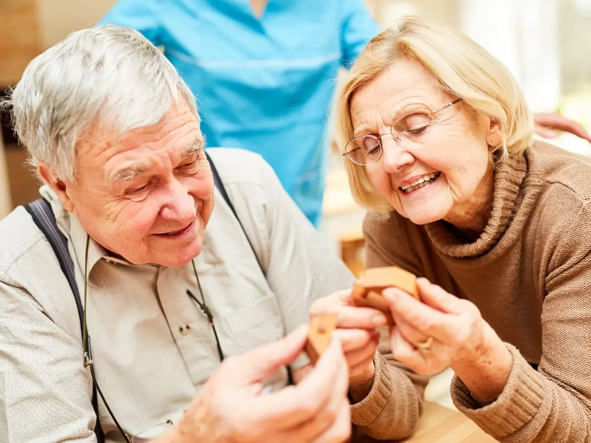 Na zdjęciu znajduje się starsza para spędzająca wspólnie czas przy aktywności związanej z układaniem klocków lub puzzli. Mężczyzna i kobieta siedzą blisko siebie przy stole, trzymając w dłoniach drewniane elementy, z zainteresowaniem i uśmiechem patrząc na nie. Atmosfera na zdjęciu jest radosna i ciepła, co sugeruje, że cieszą się chwilą spędzaną razem. Mężczyzna ma siwe włosy i nosi beżową koszulę. Na szyi ma zawieszone okulary. Kobieta ma jasne blond włosy, okulary oraz ubrana jest w brązowy sweter z golfem. W tle widoczna jest osoba ubrana w niebieski strój medyczny, która prawdopodobnie jest opiekunem lub pracownikiem domu seniora, co może wskazywać, że zdjęcie zostało zrobione w ośrodku dla osób starszych. Sceneria jest jasna, przyjemna i domowa, a na stole widać elementy układanki lub gry, co podkreśla atmosferę relaksu i wspólnego zaangażowania.