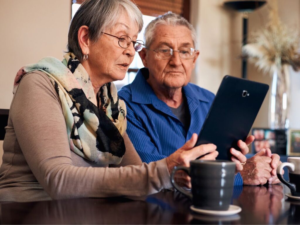 Na zdjęciu znajduje się starsza para siedząca przy stole i korzystająca z tabletu. Kobieta, z krótkimi siwymi włosami i w okularach, trzyma urządzenie w rękach i wygląda na skupioną, jakby coś czytała lub tłumaczyła. Ubrana jest w beżowy sweter, a na szyi ma kolorowy szal z delikatnym, abstrakcyjnym wzorem. Mężczyzna siedzący obok, również w okularach, patrzy na ekran tabletu z zaciekawieniem. Ma na sobie koszulę w intensywnym niebieskim kolorze, co kontrastuje z bardziej stonowanymi barwami otoczenia. Wyglądają, jakby wspólnie omawiali coś na urządzeniu – może to być rozmowa wideo, przeglądanie zdjęć lub czytanie informacji. Na stole, przed nimi, stoją dwa kubki – ciemnoszary i biały – które dodają codziennego, domowego klimatu. W tle widoczne są dekoracje wnętrza, takie jak suszone kwiaty w wazonie, lampa i zdjęcia rodzinne na półce, co sugeruje przytulne, prywatne otoczenie. Zdjęcie emanuje spokojem, bliskością i nowoczesnością w codziennym życiu osób starszych.