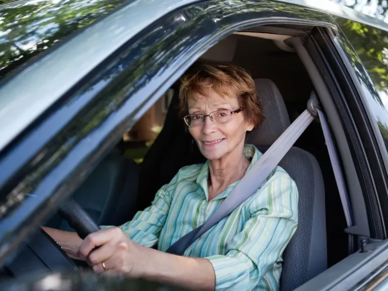 Na zdjęciu znajduje się starsza kobieta siedząca za kierownicą samochodu, widziana przez otwarte okno od strony kierowcy. Uśmiecha się serdecznie w stronę obiektywu, sprawiając wrażenie zadowolonej i pewnej siebie. Ma krótkie, jasnobrązowe włosy i nosi okulary. Ubrana jest w zieloną koszulę w paski. Na ramieniu widać zapięty pas bezpieczeństwa, co wskazuje na zachowanie zasad bezpieczeństwa podczas jazdy. Wnętrze samochodu jest widoczne w części – szare fotele i jasne obicia. W tle, za szybą, można dostrzec fragmenty zieleni, prawdopodobnie drzewa lub roślinność, co sugeruje, że zdjęcie zostało zrobione w plenerze, na terenie mieszkalnym lub w pobliżu parku. Całość emanuje spokojem i pozytywnym nastrojem, a kobieta sprawia wrażenie osoby doświadczonej i pełnej energii.