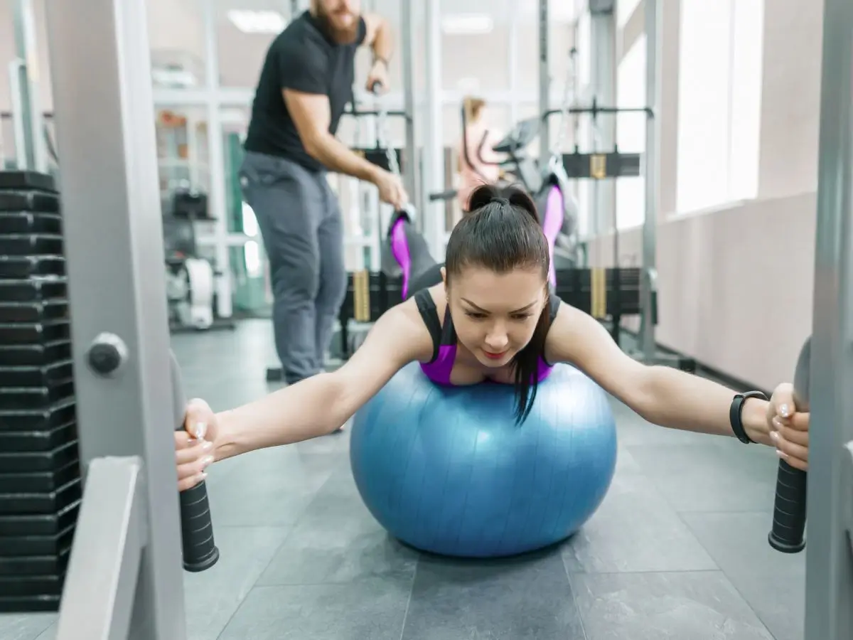 Sesja treningowa Kinesis w siłowni: Kobieta wykonuje ćwiczenie z wykorzystaniem niebieskiej piłki stabilizacyjnej, podczas gdy trener pomaga w tle. Skupienie na sile i koordynacji, a w tle widoczny sprzęt gimnastyczny
