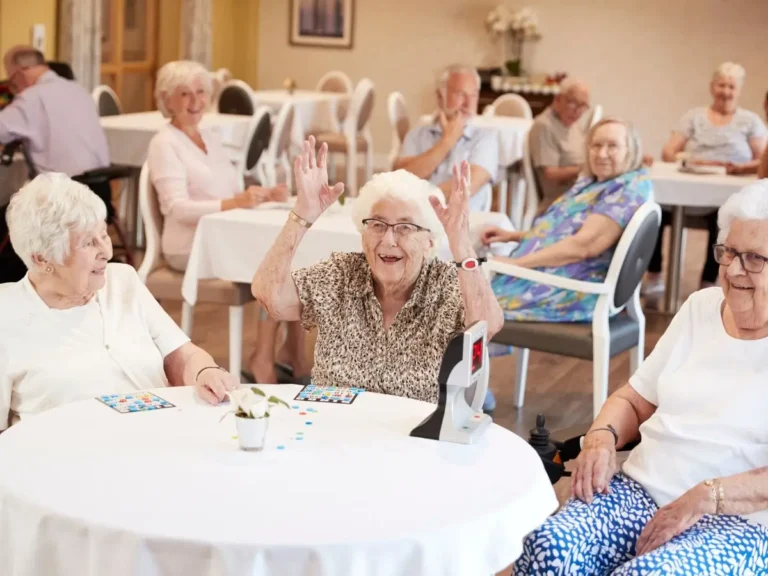Zdjęcie przedstawia grupę seniorów bawiących się w jasnej, przyjaznej przestrzeni wspólnej, prawdopodobnie w domu seniora lub ośrodku dziennej opieki. W centrum uwagi są trzy starsze kobiety siedzące przy okrągłym stole nakrytym białym obrusem, zaangażowane w grę, prawdopodobnie bingo. Szczegóły: • Na pierwszym planie: • Kobieta w środku podnosi ręce z entuzjazmem, co sugeruje, że mogła wygrać lub osiągnąć coś w grze. Uśmiecha się szeroko i wygląda na zadowoloną. • Po jej lewej i prawej stronie siedzą dwie inne kobiety, które również są uśmiechnięte i w dobrym nastroju. • Na stole widoczne są plansze do gry, drobne żetony oraz mała doniczka z zieloną rośliną jako ozdoba. • Na stole znajduje się też elektroniczne urządzenie z wyświetlaczem, które prawdopodobnie jest częścią gry. • W tle: • Przy innych stołach siedzą inni seniorzy, zajęci rozmowami lub obserwowaniem gry. Są uśmiechnięci i wyglądają na zrelaksowanych. • Na jednej ze ścian widoczna jest dekoracja, a w rogu stoi komoda z kwiatami, co nadaje pomieszczeniu domowego charakteru. Atmosfera: Zdjęcie pokazuje ciepłą, radosną atmosferę. Seniorzy wyglądają na zaangażowanych, a otoczenie jest jasne, czyste i przytulne. To miejsce, które sprzyja integracji i aktywnemu spędzaniu czasu.