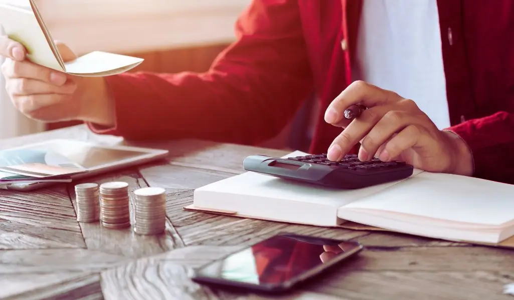 Na zdjęciu widać osobę ubraną w czerwony sweter, która liczy swoje finanse. Jedną ręką trzyma otwarty notatnik lub książeczkę czekową, a drugą wciska przyciski kalkulatora, robiąc obliczenia. Na drewnianym stole leży stos monet, co symbolizuje oszczędzanie lub planowanie budżetu. Obok znajduje się telefon oraz tablet, co sugeruje korzystanie z nowoczesnych narzędzi finansowych. Zdjęcie oddaje atmosferę skupienia, odpowiedzialności i zarządzania pieniędzmi – może odnosić się do tego, ile zarabia osoba z orzeczeniem i jak planuje swoje wydatki.