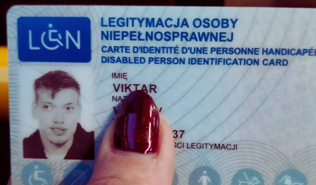 Zdjęcie przedstawia Legitymację osoby niepełnosprawnej – oficjalny dokument potwierdzający status osoby z niepełnosprawnością. Karta ma niebieskie logo z symbolem wózka inwalidzkiego i napisy w trzech językach. Widoczne jest czarno-białe zdjęcie mężczyzny o imieniu Viktar oraz częściowo zasłonięte nazwisko. Dokument trzyma dłoń z czerwonym, błyszczącym paznokciem, co kontrastuje z niebiesko-białą kolorystyką karty. Zdjęcie ma osobisty charakter, ukazując zarówno formalność dokumentu, jak i codzienny aspekt jego użytkowania.