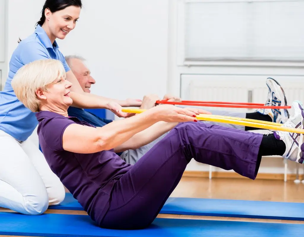 Zdjęcie przedstawia grupową sesję ćwiczeń rehabilitacyjnych lub fizjoterapeutycznych, w której uczestniczą dwie osoby starsze – kobieta i mężczyzna – pod opieką młodej instruktorki lub fizjoterapeutki. W pierwszym planie znajduje się starsza kobieta z krótkimi, blond włosami, ubrana w ciemnofioletowy strój sportowy. Siedzi na niebieskiej macie do ćwiczeń w pozycji półleżącej, z kolanami zgiętymi i stopami uniesionymi. Trzyma w dłoniach żółtą taśmę oporową, którą zahaczyła o stopy, wykonując ćwiczenie wzmacniające mięśnie brzucha i pleców. Za nią znajduje się starszy mężczyzna w niebieskiej koszulce i szarych spodniach, również trzymający taśmę (w kolorze czerwonym) i wykonujący to samo ćwiczenie. Z lewej strony klęczy młoda kobieta w niebieskim stroju medycznym, najprawdopodobniej instruktorka lub rehabilitantka. Uśmiecha się i wspiera ćwiczących, utrzymując kontakt wzrokowy i prawdopodobnie korygując postawę. Tło to jasne, czyste pomieszczenie z białymi ścianami, dużym oknem z roletą i grzejnikiem. Podłoga jest wykonana z jasnego drewna. Całość emanuje pozytywną energią, zaangażowaniem i atmosferą troski o zdrowie.