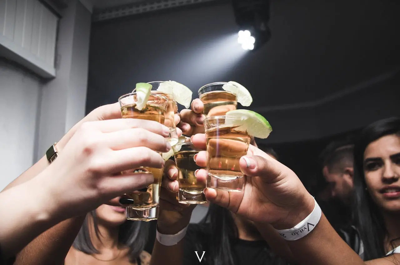 Zdjęcie przedstawia grupę osób wznoszących toast przy pomocy kieliszków z bursztynowym alkoholem – najprawdopodobniej tequilą – każdy kieliszek ozdobiony jest plasterkiem limonki na rancie. W centrum kadru znajduje się sześć zbliżonych do siebie rąk trzymających przezroczyste kieliszki typu shot. Szkło wypełnione jest jasnozłotym trunkiem, który w połączeniu z cytrusowym dodatkiem sugeruje klasyczne podanie tequili. Ręce należą do osób o różnej karnacji – widać zarówno kobiece dłonie z ciemnym lakierem na paznokciach, jak i męskie, z opaskami na nadgarstkach, co może sugerować uczestnictwo w imprezie klubowej lub zamkniętym evencie. Tło jest lekko rozmazane – widać reflektory klubowe i sylwetki osób, w tym kobietę po prawej stronie patrzącą w stronę toastu. Atmosfera zdjęcia oddaje klimat nocnego życia – celebrację, radość, wspólne świętowanie.