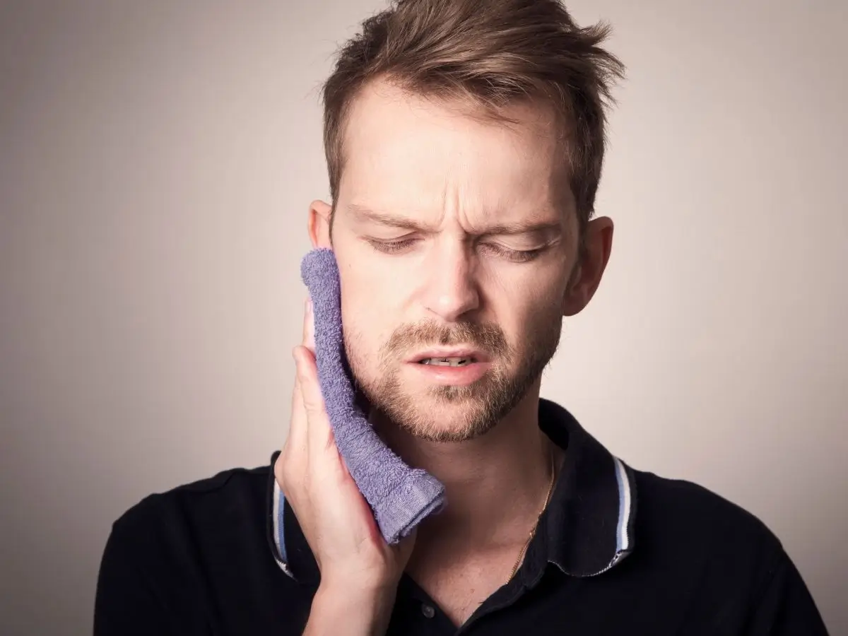 Mężczyzna z grymasem bólu na twarzy przykłada ręcznik do policzka, wyraźnie odczuwając dyskomfort po stronie szczęki. Obraz ilustruje stan po wyrwaniu zęba i podkreśla potrzebę odpowiedniej troski o proces gojenia, w tym unikania alkoholu po zabiegu stomatologicznym
