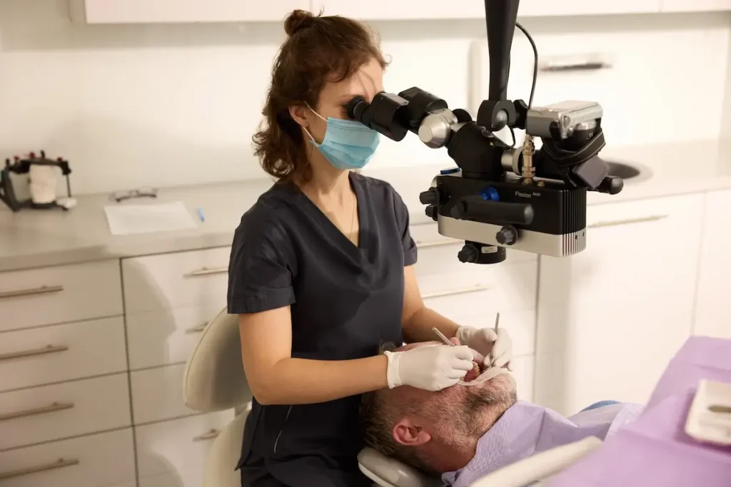 Dentystka w maseczce i rękawiczkach wykonuje precyzyjny zabieg stomatologiczny u leżącego pacjenta, korzystając z mikroskopu zabiegowego. Nowoczesne, sterylne wnętrze gabinetu podkreśla profesjonalizm i zaawansowaną technologię kliniki