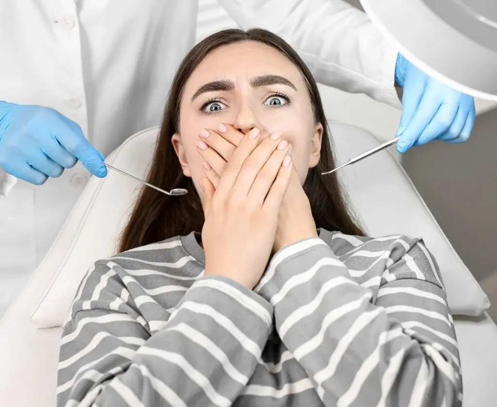 Przerażona młoda kobieta siedzi na fotelu dentystycznym i zakrywa usta dłońmi. Za nią widoczne są ręce stomatologa w niebieskich rękawiczkach, trzymającego narzędzia dentystyczne. Obraz ilustruje silny lęk przed wizytą u dentysty i potrzebę przełamania strachu w trosce o zdrowie jamy ustnej.